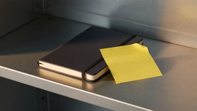 A closed notebook and a blank sticky note on a steel shelf, warm side light, no labels and no logos.