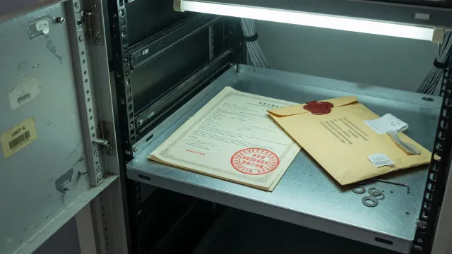 An open server-rack door, a stamped paper certificate, and a sealed envelope on a steel shelf, cool room light, no logos.
