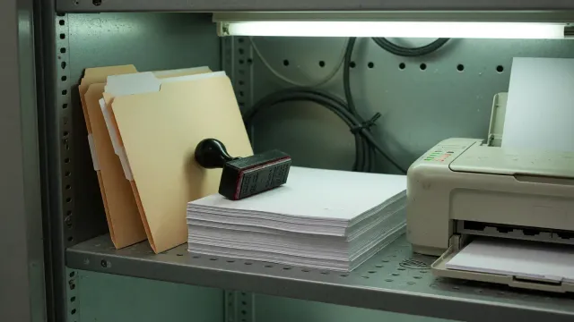 A server-closet shelf with two manila folders, a rubber stamp, blank paper, and a small printer under fluorescent light.