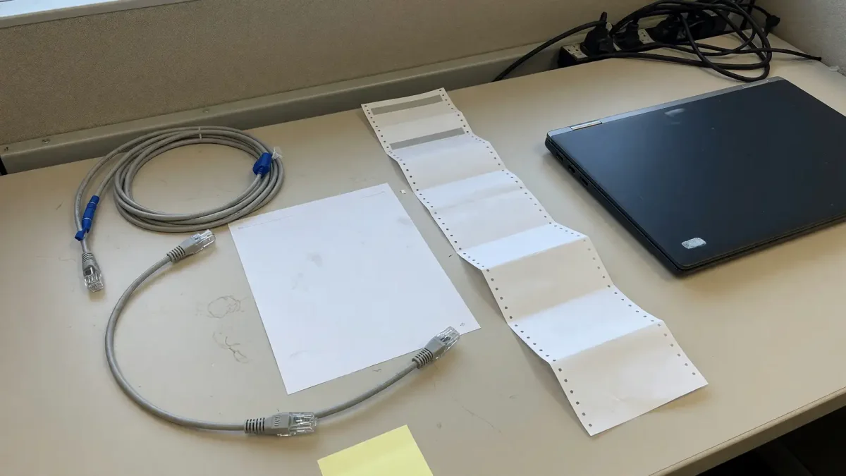 Two blank paper printouts, one short and one long, beside unplugged ethernet cables and a closed laptop on a beige desk.