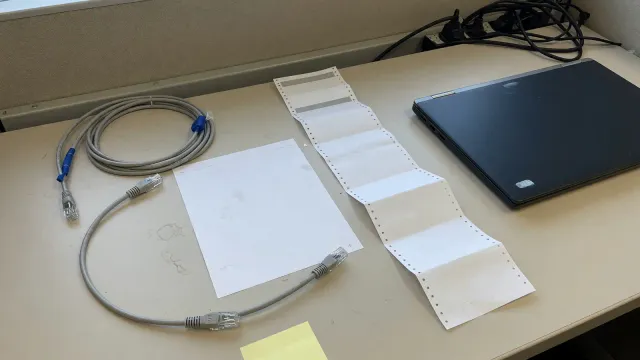 Two blank paper printouts, one short and one long, beside unplugged ethernet cables and a closed laptop on a beige desk.
