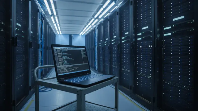 Laptop in a server aisle showing a terminal with a cluster upgrade command, cool lighting, no readable project names or logos.