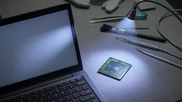 An open laptop on a bench beside a small TPM chip on an anti-static mat, cool shop light, no vendor logos and no readable text on the screen.